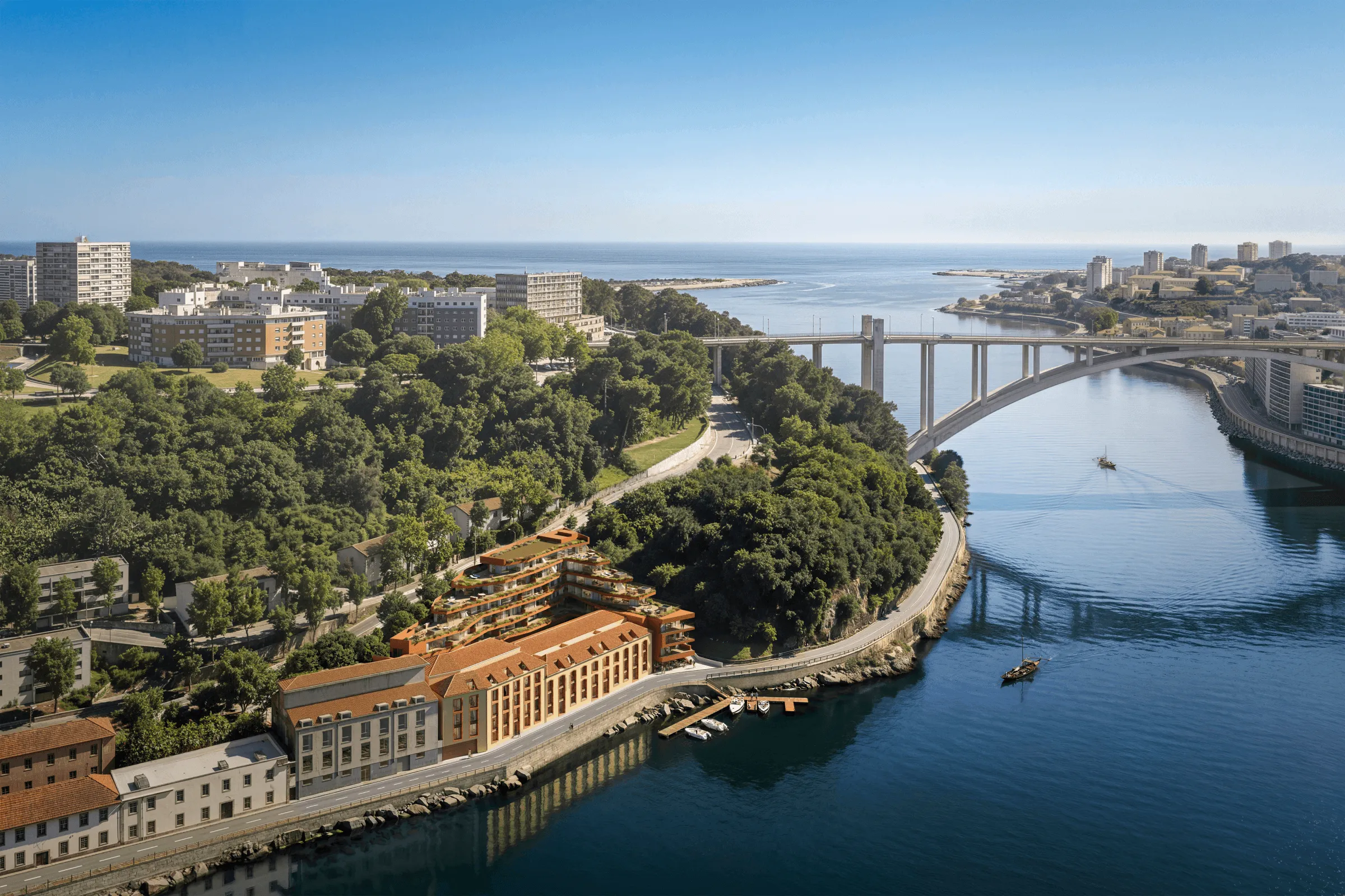 Panoramic aerial view of a coastal city, featuring lush green areas and residential buildings on the left, a large bridge spanning a waterway in the center, and the sea in the background under a clear sky.